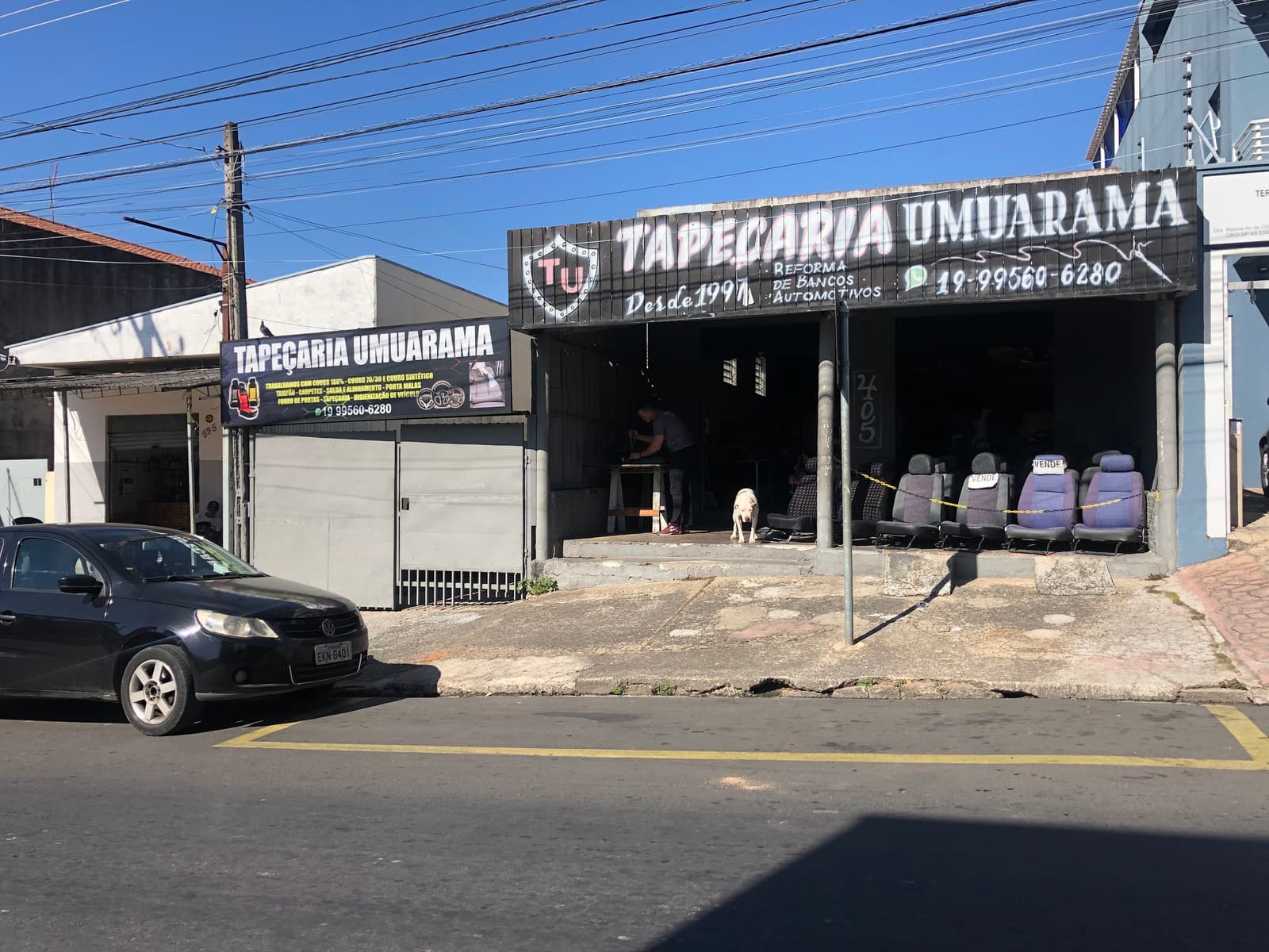 Fachada da Tapeçaria Umuarama durante o dia, com a placa pintada 'Desde 1997 — Reforma de bancos automotivos' e bancos expostos na entrada